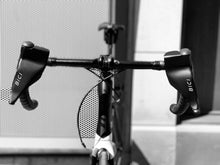 Load image into Gallery viewer, A head on shot of a high end road bike&#39;s handlebars showing two Bici Shield winter cycling hand protectors attached to both brake levers.