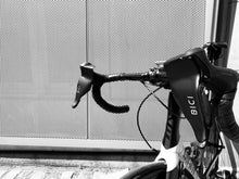 Load image into Gallery viewer, The handlebars of a white roadbike with black handlebars with two bici shields attached to its brake levers, photographed in front of a metal mesh wall.