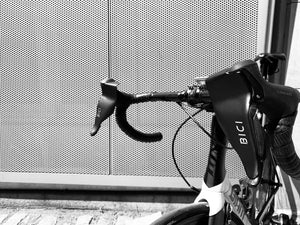 The handlebars of a white roadbike with black handlebars with two bici shields attached to its brake levers, photographed in front of a metal mesh wall.